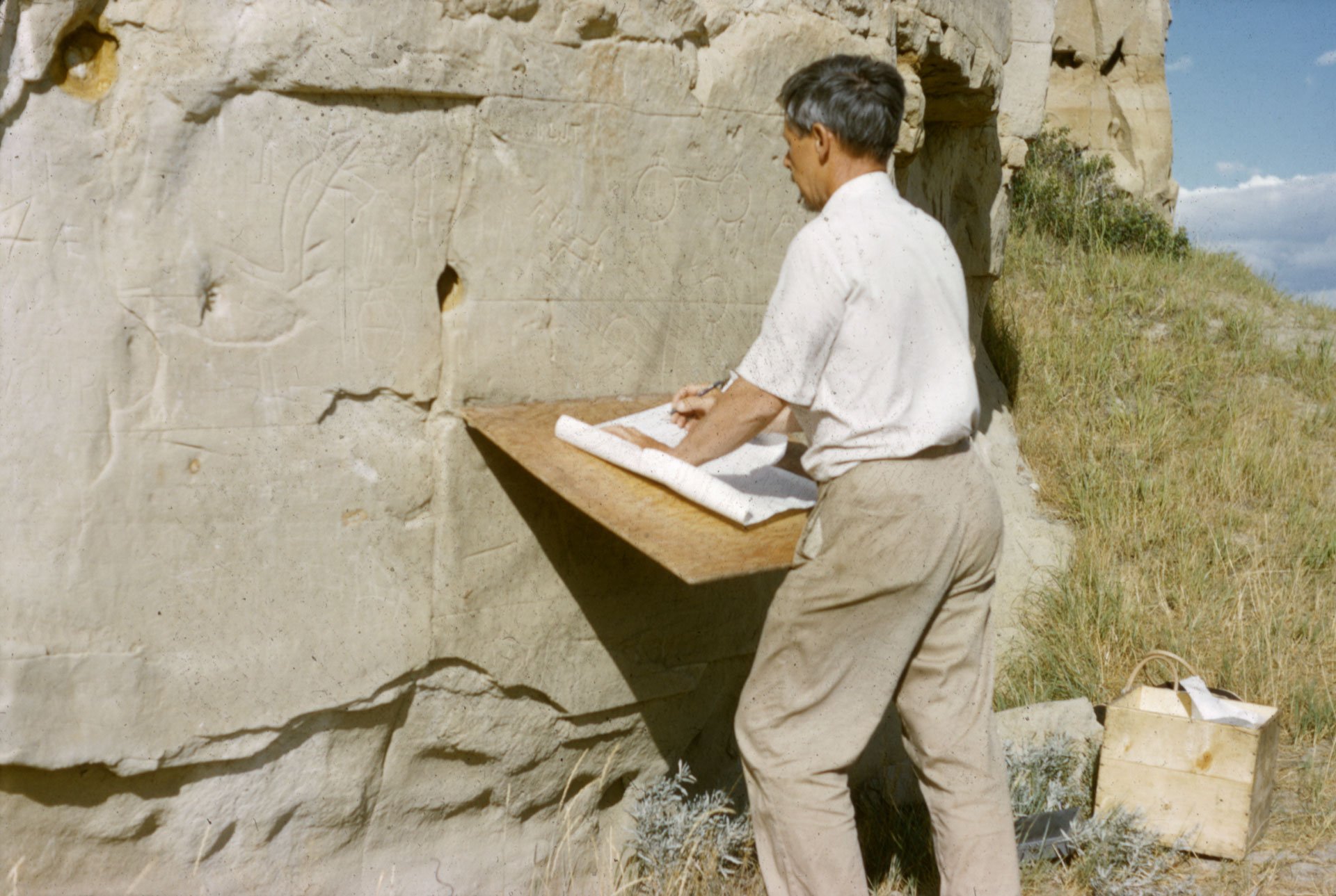 Picture of Selwyn Dewdney checking tracings of petroglyphs at Áísínai'pi, in August 1960