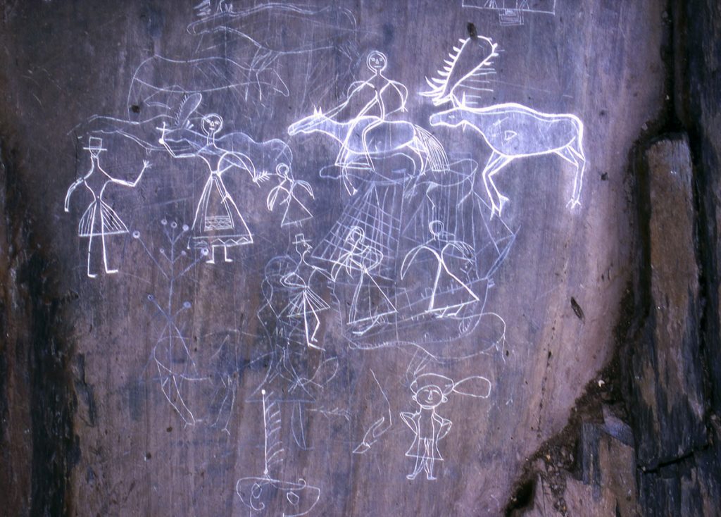 McGowan Lake, Nova Scotia. Carving of a hunting scene where a Mi'kmaq man, smoking a pipe and wearing a hat, sends two dogs to hunt moose.