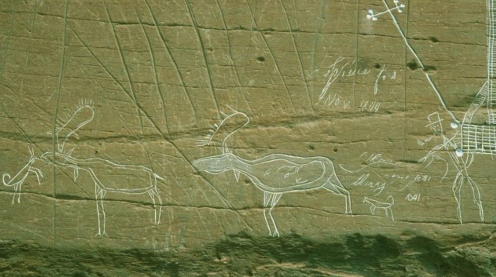 McGowan Lake, Nova Scotia. Carving of a hunting scene where a Mi'kmaq man, smoking a pipe and wearing a hat, sends two dogs to hunt moose.