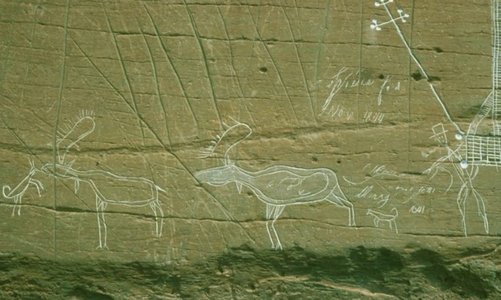 McGowan Lake, Nova Scotia. Carving of a hunting scene where a Mi'kmaq man, smoking a pipe and wearing a hat, sends two dogs to hunt moose.
