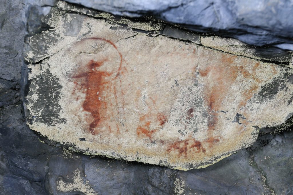 Photographie présentant différents personnages, soit un anthropomorphe, qui semble exécuter une danse muni d’un cerceau orné de fils, se tient devant un être cornu (à droite) accompagné d’un serpent (tenu à la main).
