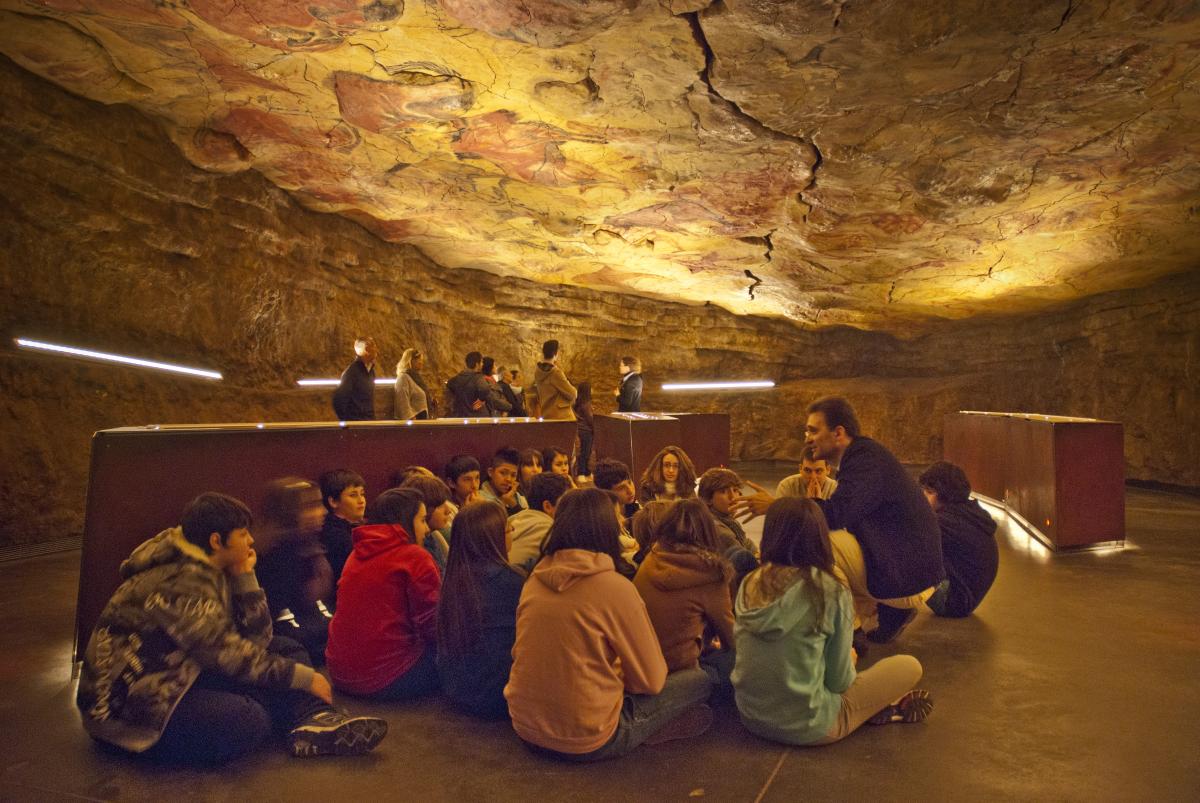 A group of young visitors is sitting in a cave and listening to a teacher, in a cave where we see several bulls painted on the ceiling.